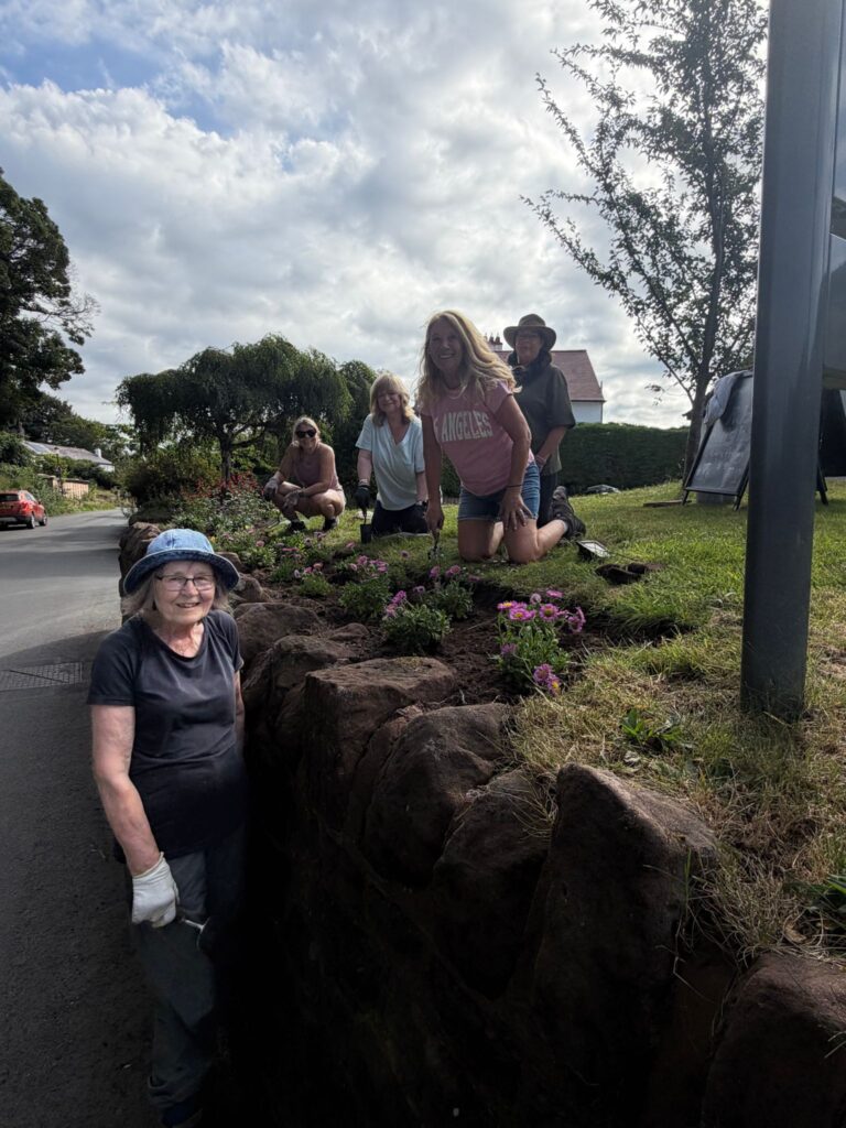 Five ladies in the middle of planting in the border outside GVH, smiling at the camera.